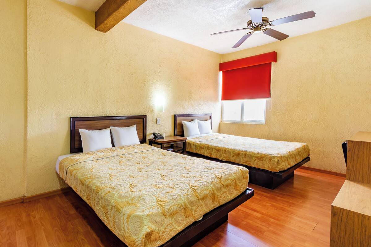 A hotel room with two beds, wooden flooring, a ceiling fan, and a window with red curtains.