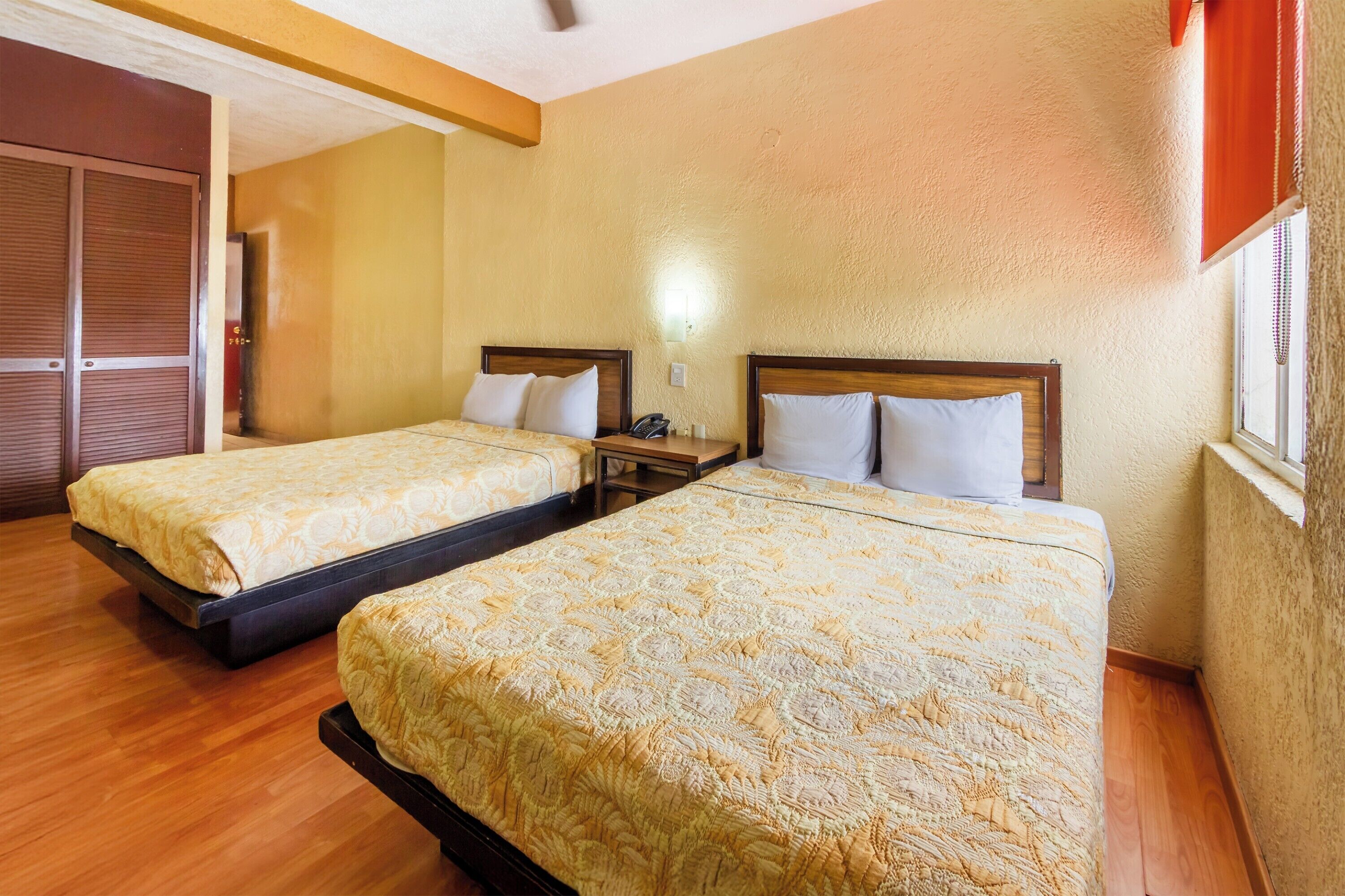 A hotel room with two beds, wooden flooring, a wardrobe, and a window with blinds.