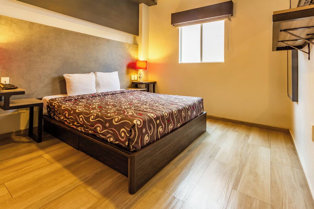 A modern bedroom with a large bed, wooden flooring, a nightstand with a lamp, and a window providing natural light.