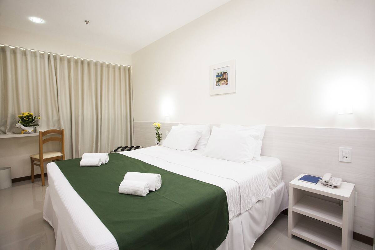 A hotel room with a neatly made bed, a bedside table with a phone and a book, a chair, and a small table with a vase of flowers.