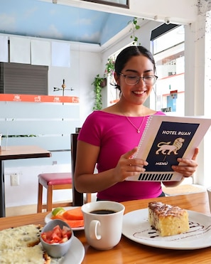 Una persona leyendo un menú en una mesa, con una porción de pastel, una taza de café y un bol de fruta.