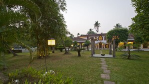 A courtyard with a pathway leading to a building with a tiled roof and arched windows. - Purity at Lake Vembanad (Cherthala)