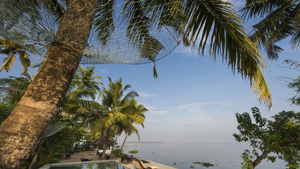 A poolside area with lounge chairs, palm trees, and a view of the sea. - Purity at Lake Vembanad (Cherthala)