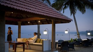 A beachside lounge with a covered seating area, palm trees, and a view of the sea. - Purity at Lake Vembanad (Cherthala)