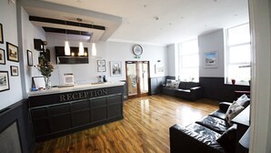 A reception area with a wooden floor, a reception desk labeled "RECEPTION," a clock on the wall, and a seating area with a sofa and a chair. - Lansdowne Hotel (Hastings)
