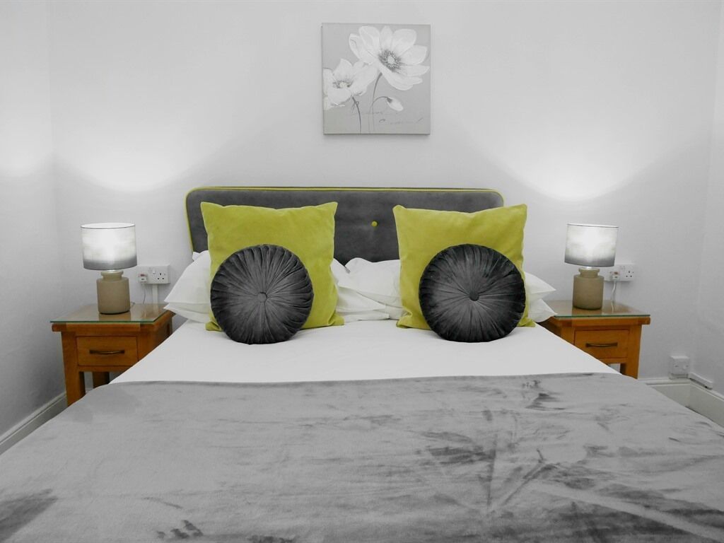 A bed with a grey headboard, two green pillows, and a grey bedspread. There are bedside lamps on either side of the bed.