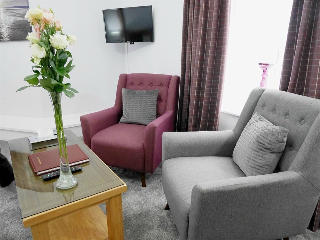 Una sala de estar con un sofá gris, un sillón rojo, una mesa de centro de vidrio con un jarrón de flores, un televisor de pantalla plana montado en la pared y una ventana con cortinas.