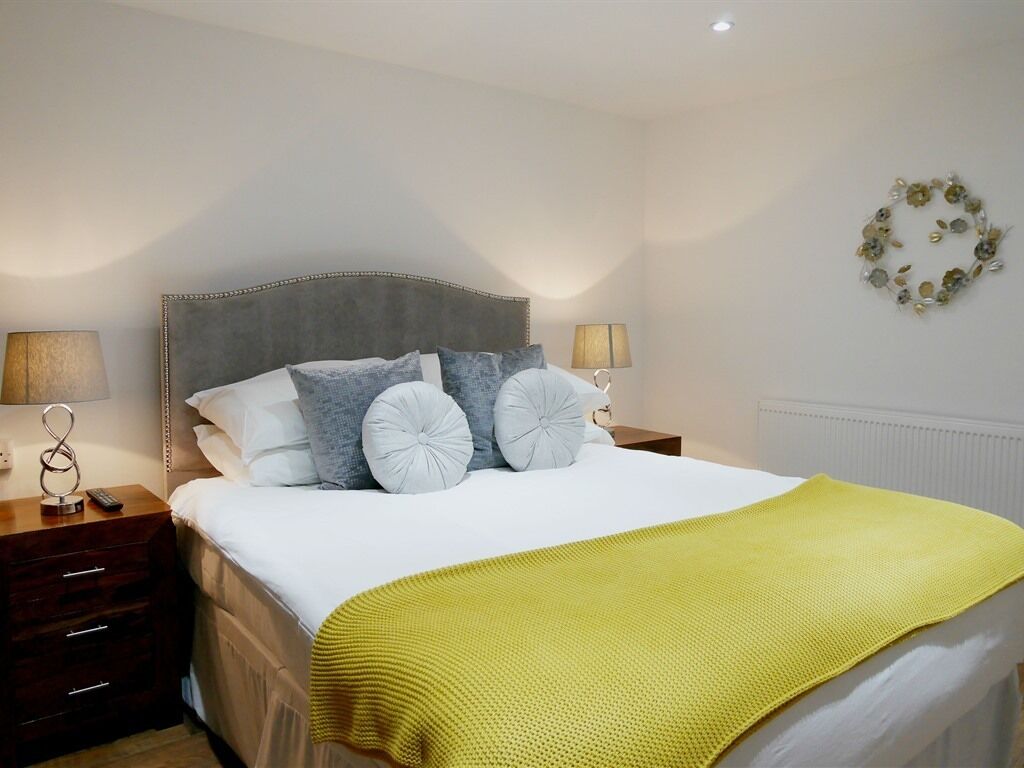 A neatly made bed with a white duvet and a mustard yellow throw blanket. There are two bedside tables with lamps and a decorative wall piece.
