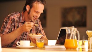 Une personne est assise à une table, devant un ordinateur portable, et prend son petit-déjeuner : elle mange des céréales, boit du café et du jus d'orange.