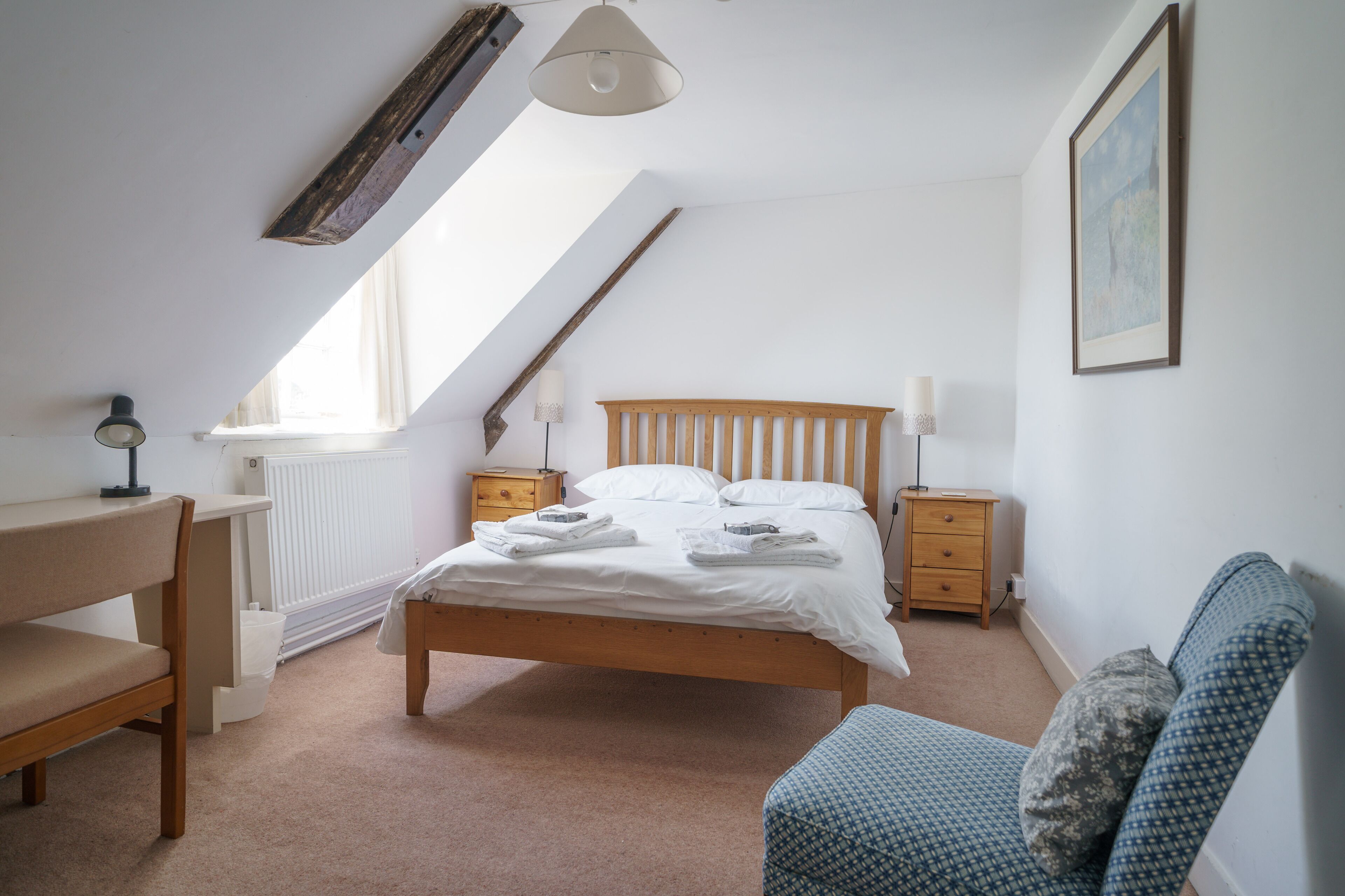 Un dormitorio con techo inclinado, una cama con ropa blanca, cabecera de madera, mesita de noche con lámpara, una silla e imagen enmarcada en la pared.