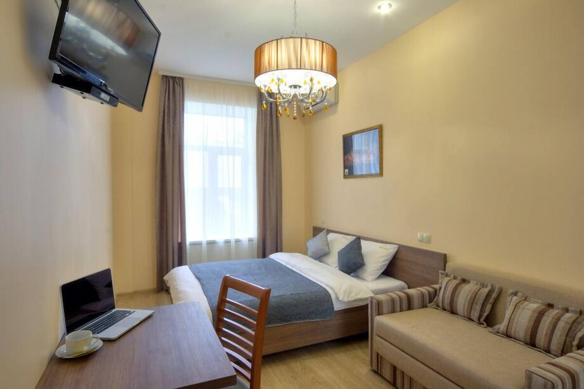 A hotel room with a bed, a desk with a laptop, a chair, a sofa, a television, and a chandelier.