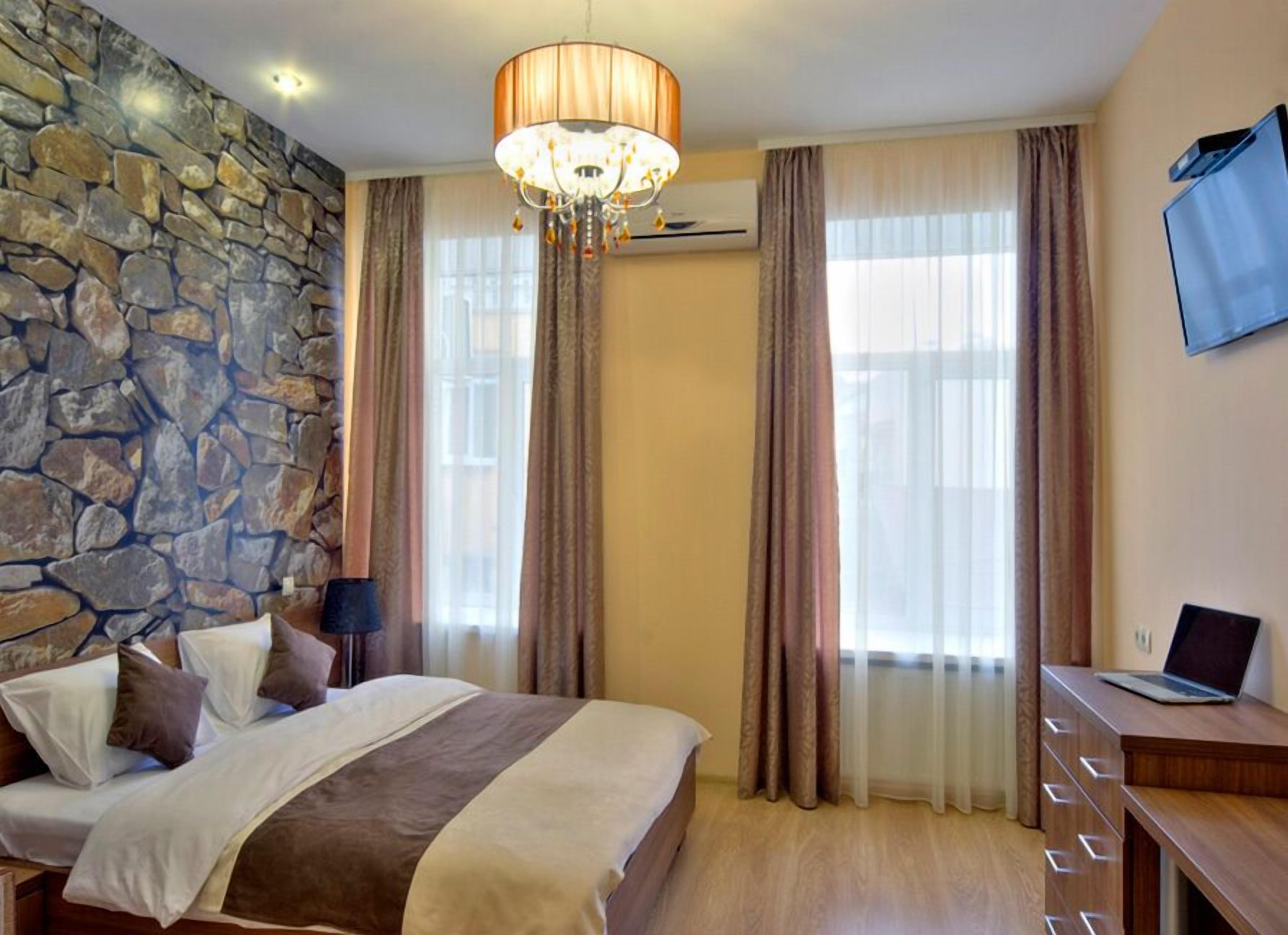 A bedroom with a stone wall, a bed with pillows, a desk with a laptop, and a chandelier.