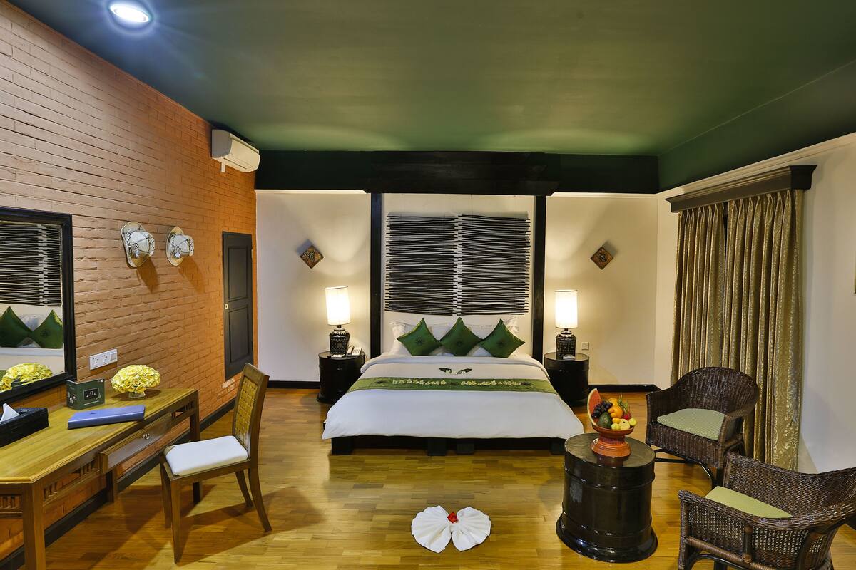 Quarto de hotel com escrivaninha de madeira, cama com colcha verde e branca, duas mesas de cabeceira com abajures, uma cadeira de vime e uma janela grande com persianas.