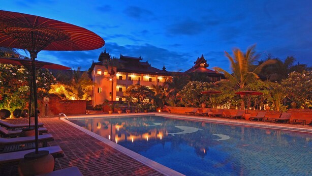 A swimming pool with lounge chairs and umbrellas, surrounded by tropical plants and a building with traditional architecture. - Amazing Bagan Resort (Nyaung-U)
