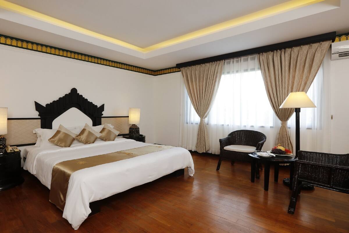Quarto de hotel com uma cama grande, duas mesas de cabeceira com luminárias, uma cadeira, uma mesinha com frutas e uma janela com cortinas.