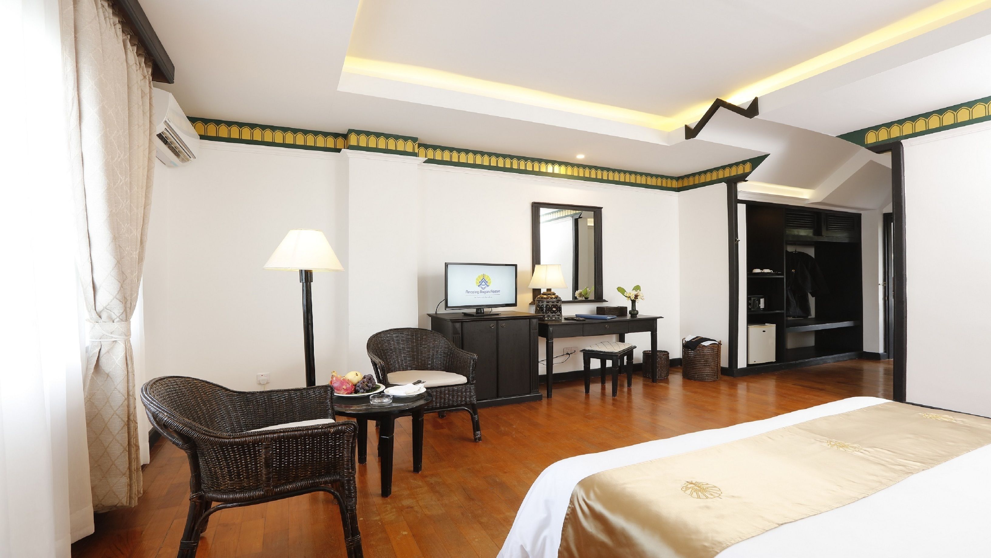 A hotel room with a bed, a desk with a computer, a chair, a lamp, and a small table with fruits.