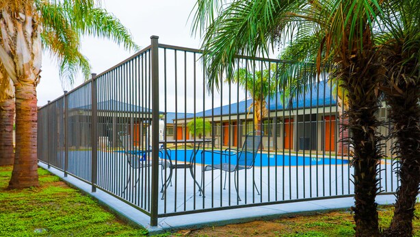 Uma área de piscina cercada, com piscina azul, palmeiras e um edifício com telhado azul ao fundo.