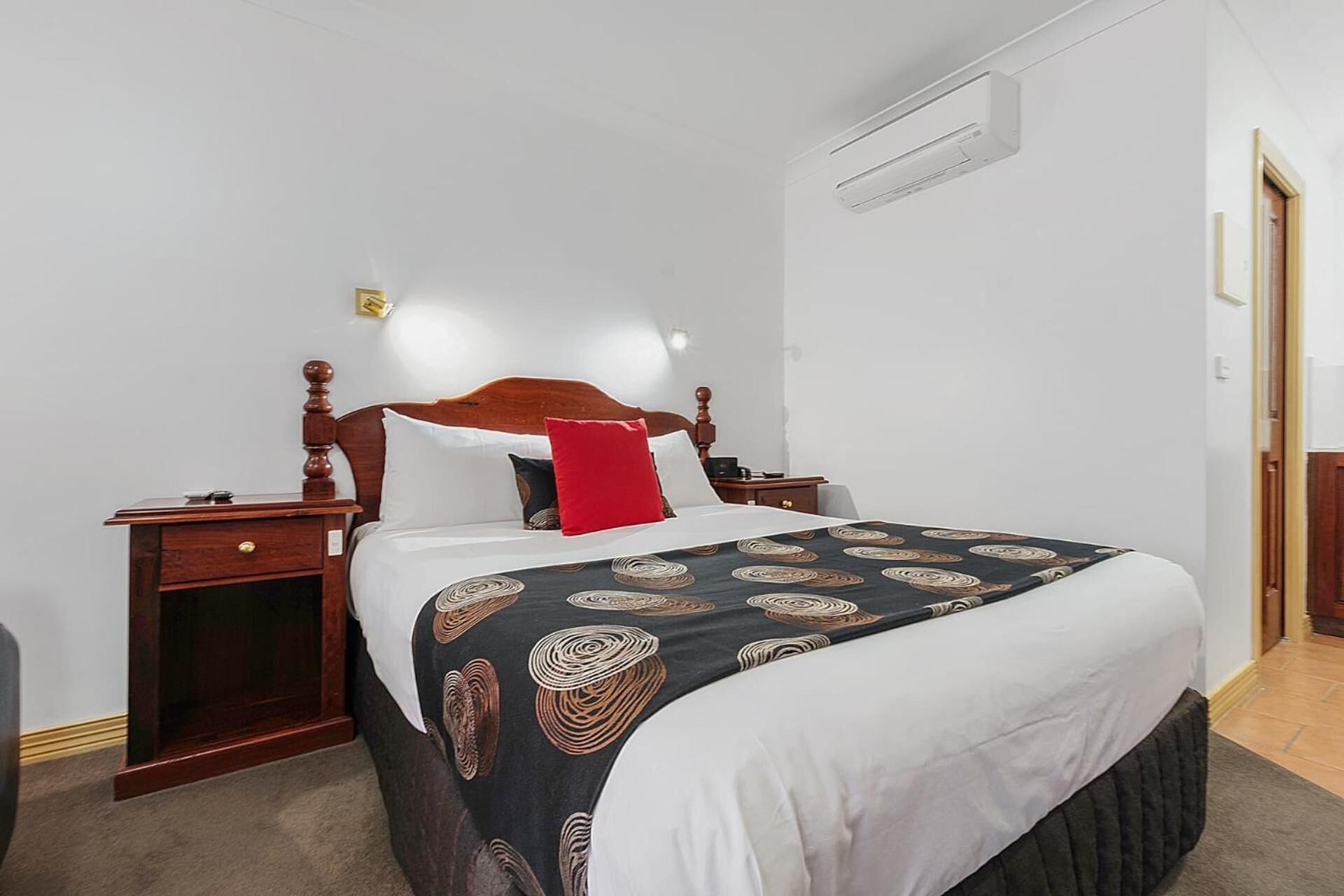 Une chambre d’hôtel avec un cadre de lit en bois, une couvre-lit à motifs foncés, une couette blanche et un oreiller rouge. On y trouve une table de chevet en bois avec une lampe, un climatiseur fixé au mur et une porte sur le côté droit.