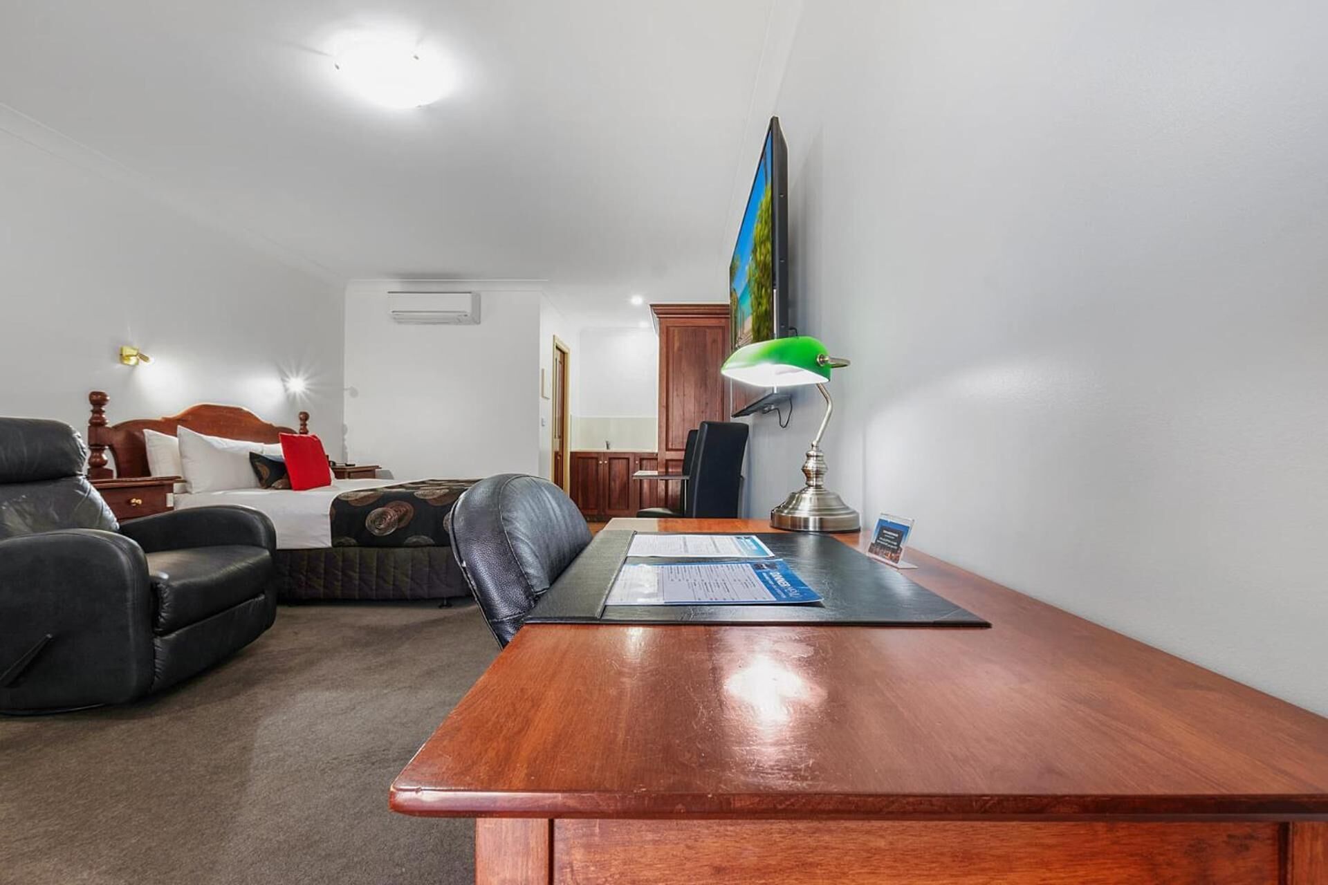 Quarto de hotel com escrivaninha de madeira, poltrona de couro, cama com travesseiro vermelho e abajur verde.