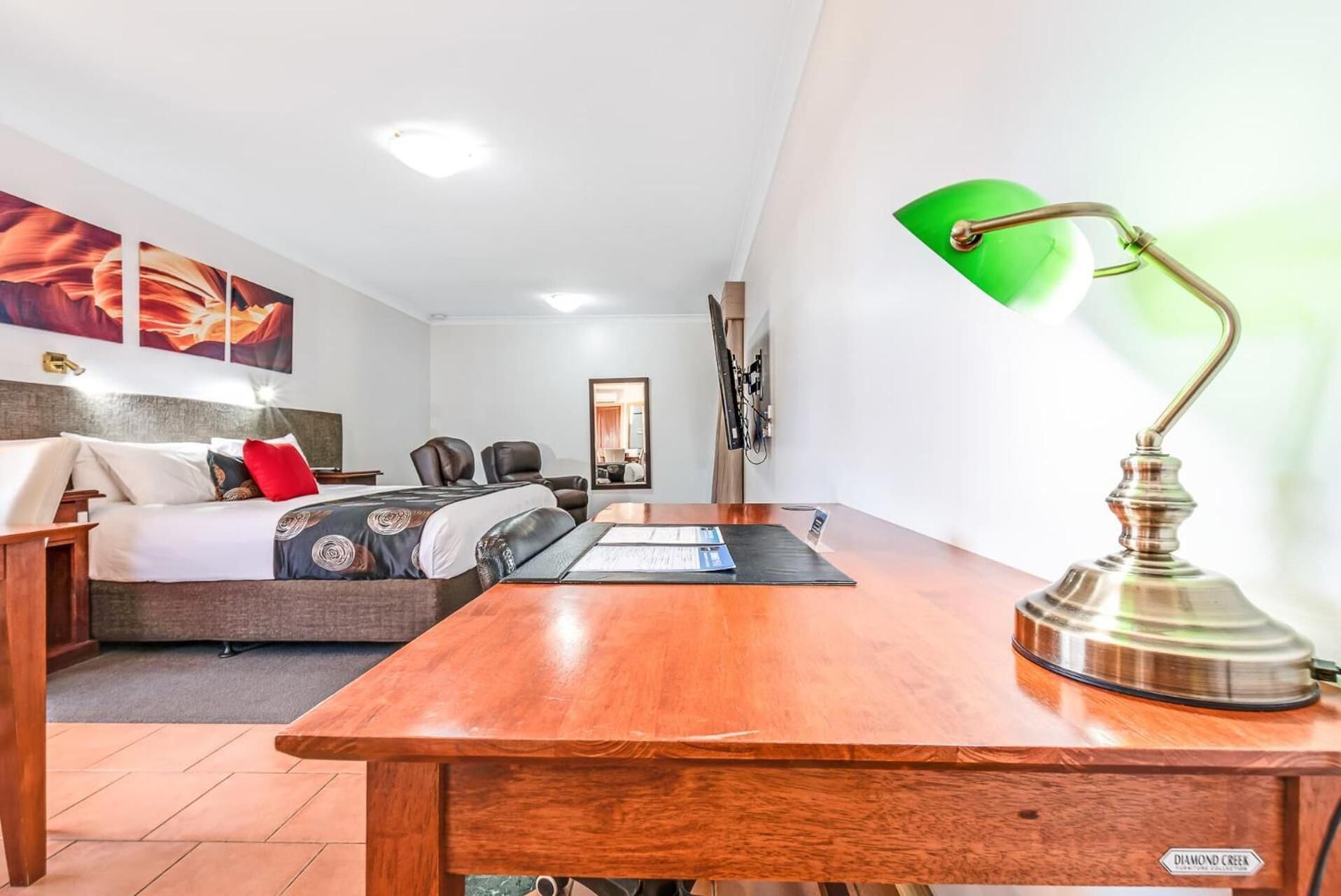 A hotel room with a bed, a desk, a green desk lamp, and artwork on the wall.