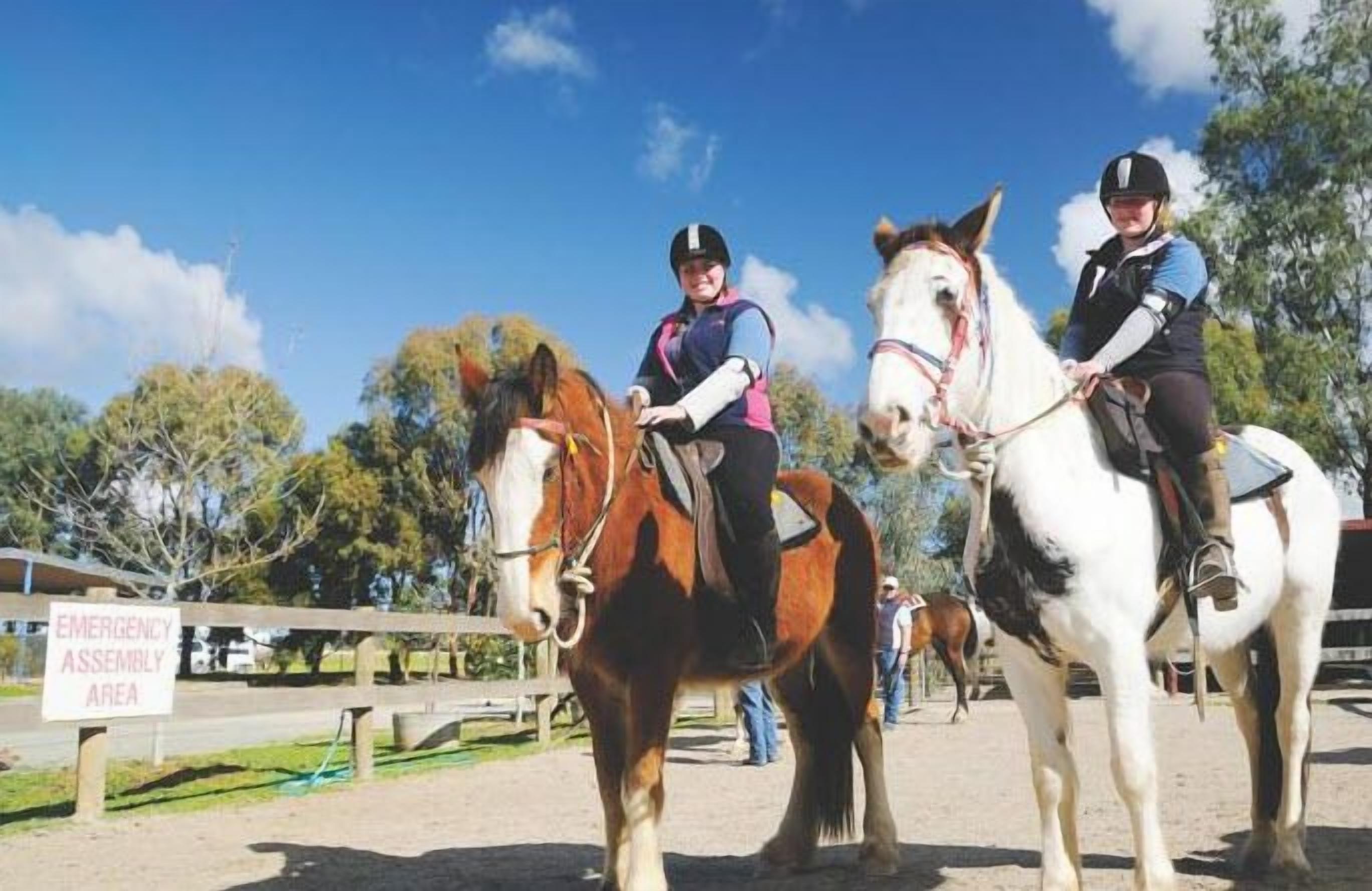 Tres personas montando a caballo en un espacio al aire libre con un letrero que dice "EMERGENCY ASSEMBLY AREA".