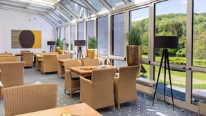 Un restaurant avec des meubles en osier, une grande fenêtre donnant sur un paysage verdoyant et une table dressée.