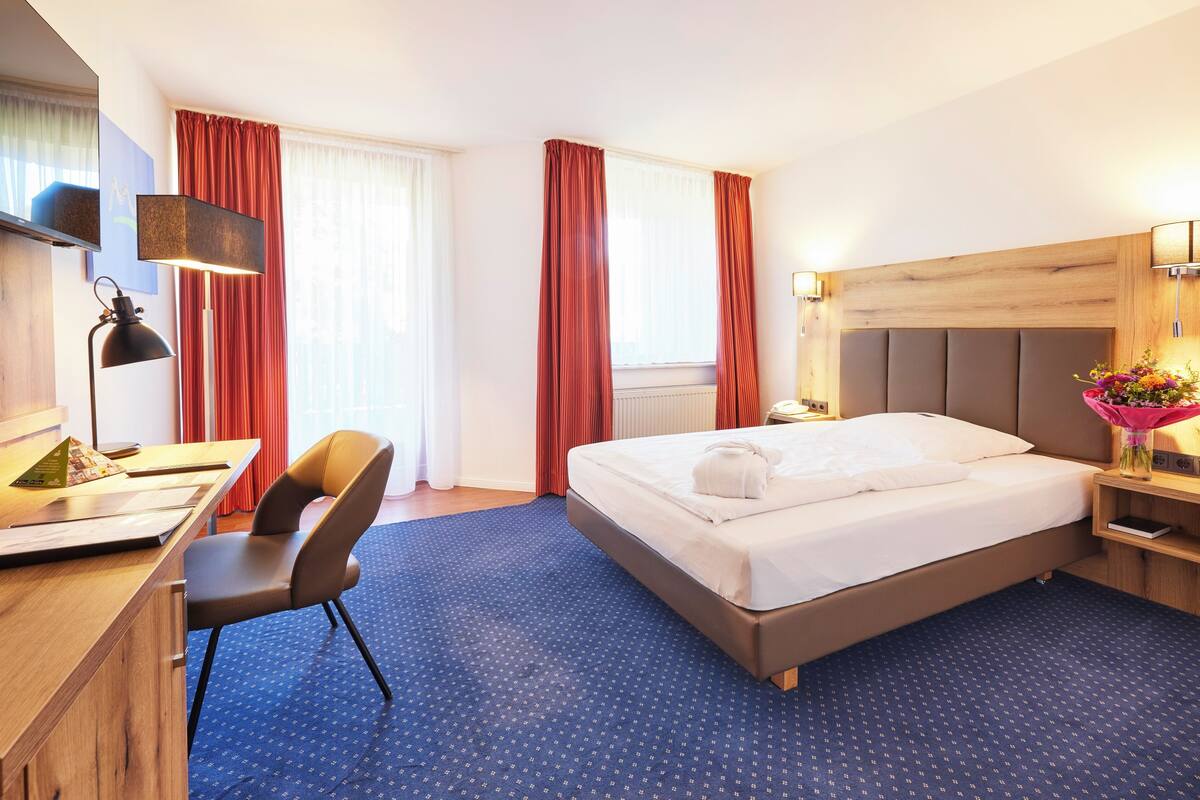 A hotel room with a bed, desk, chair, and a large window with curtains.