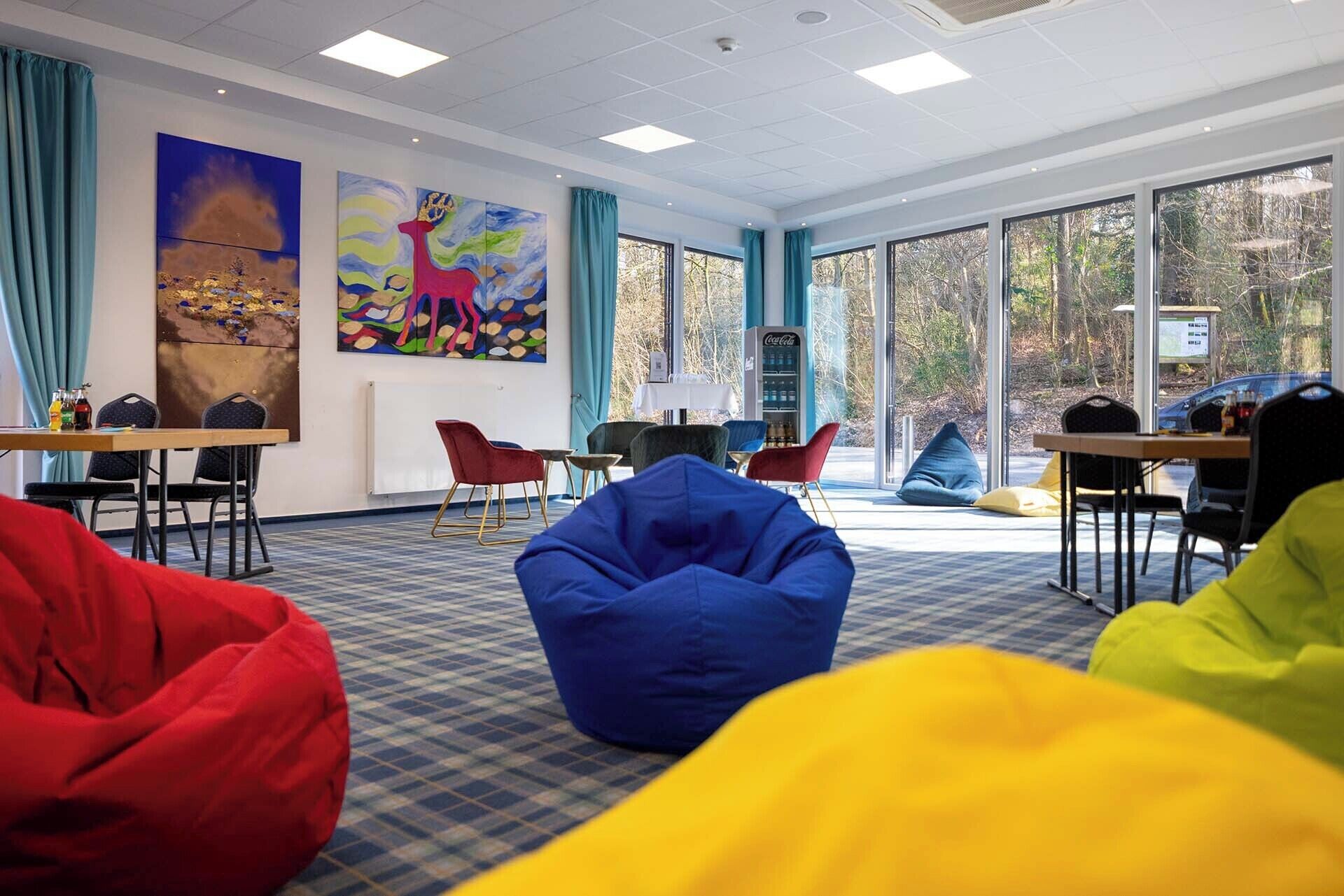 Ein modernes Zimmer mit großen Fenstern, bunten Sitzsäcken und Kunstwerken an den Wänden.