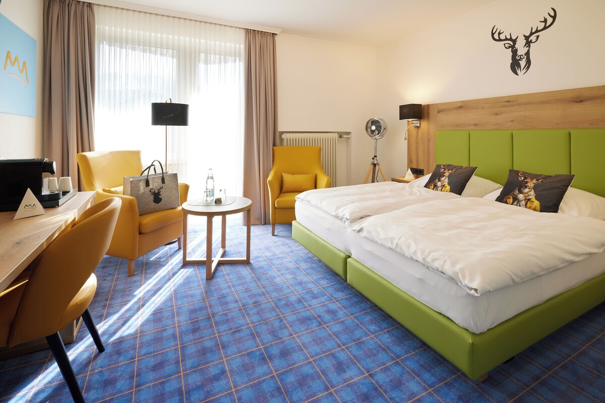 A hotel room with a bed, two yellow chairs, a desk with a laptop, a small table with a lamp, and a radiator.
