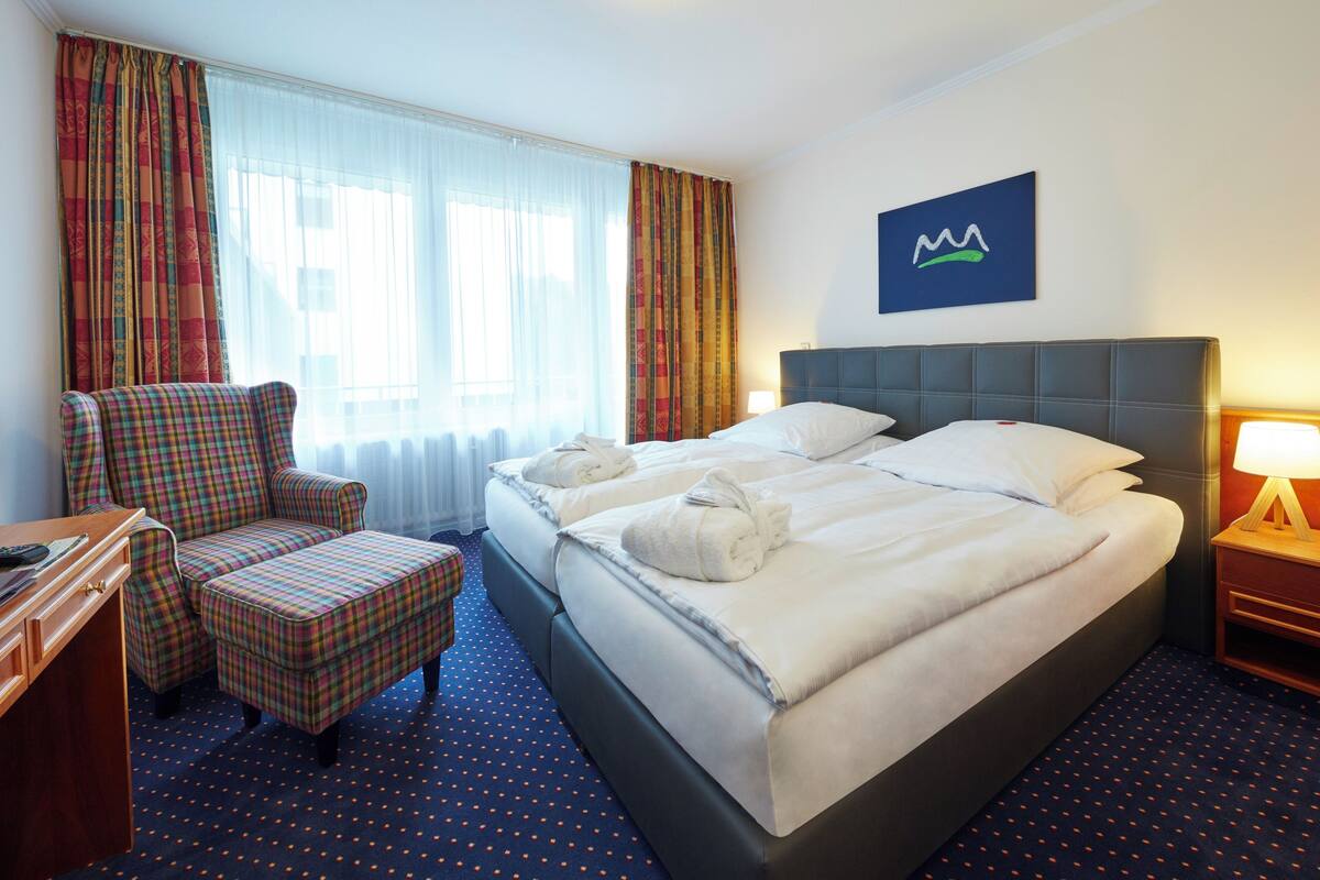 A hotel room with a large bed, a chair, a desk, and a window with curtains.