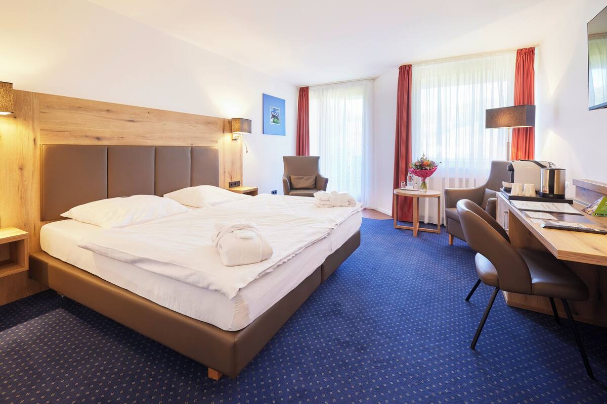 A hotel room with a large bed, a desk with a chair, a small table with a vase, and a window with curtains.