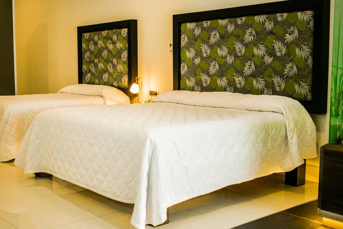 A double bed with a quilted white duvet, two bedside tables with lamps, and a wall with a leaf pattern.