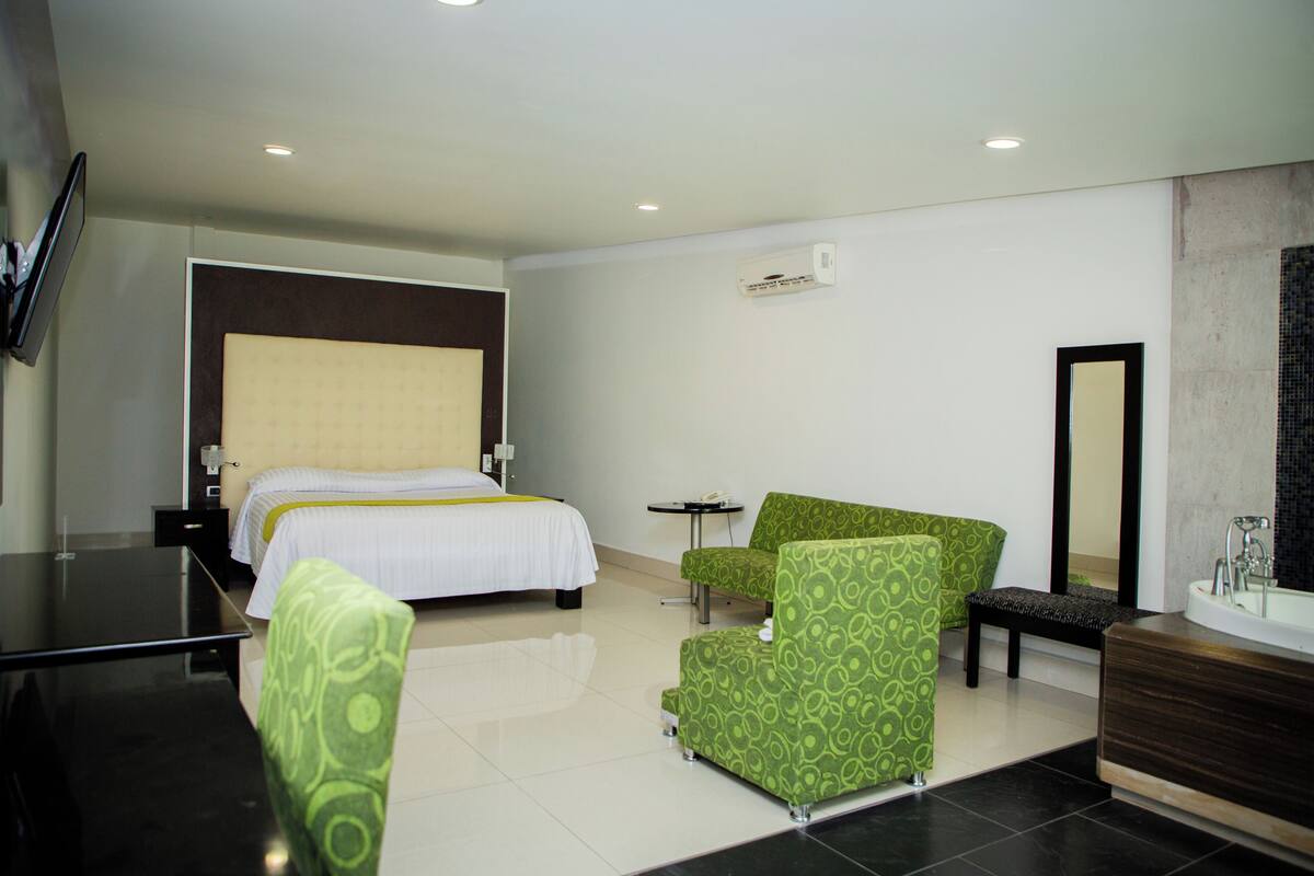 A hotel room with a bed, a green armchair, a small table, a bench, a mirror, and a bathroom with a sink and a bathtub.