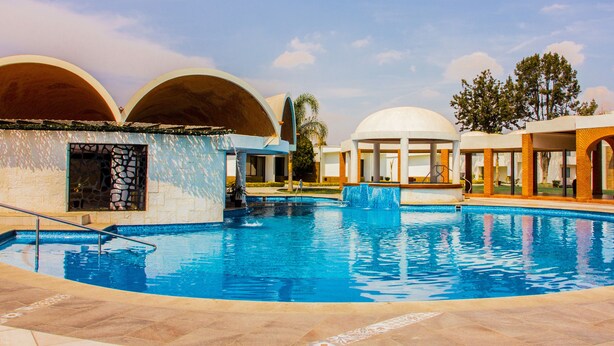 A swimming pool with a tiled deck, surrounded by buildings with arched structures and a clear sky.