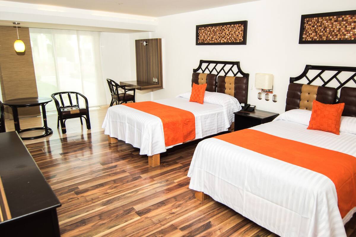 A hotel room with two beds, wooden flooring, a desk, chairs, and a small table.