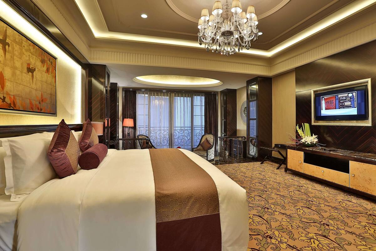 A hotel room with a large bed, a TV, a chandelier, and a view of the outside through the window.
