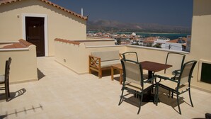 Eine Terrasse mit Tisch und Stühlen, einer Tür und Blick auf eine Küstenstadt und Berge.