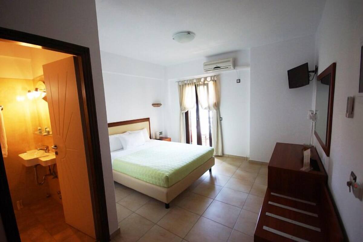 A hotel room with a bed, a small bedside table, a TV mounted on the wall, and a window with curtains.