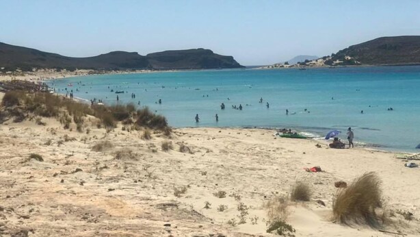 Ein Strand mit klarem, blauem Wasser, Sanddünen und sanften Hügeln im Hintergrund.