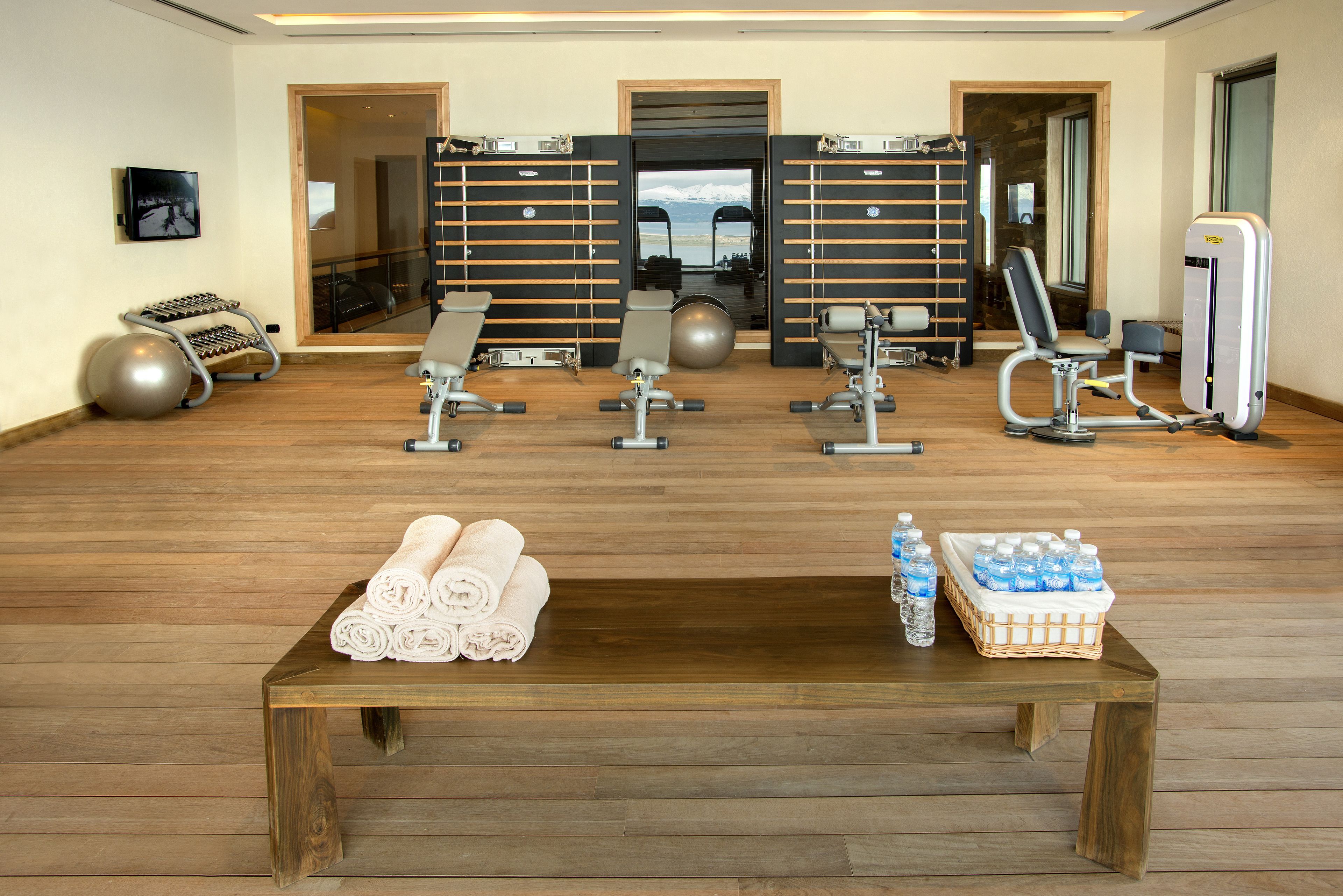 Academia com equipamentos de ginástica, piso de madeira, mesa com toalhas e garrafas de água, e vista para montanhas pela porta.