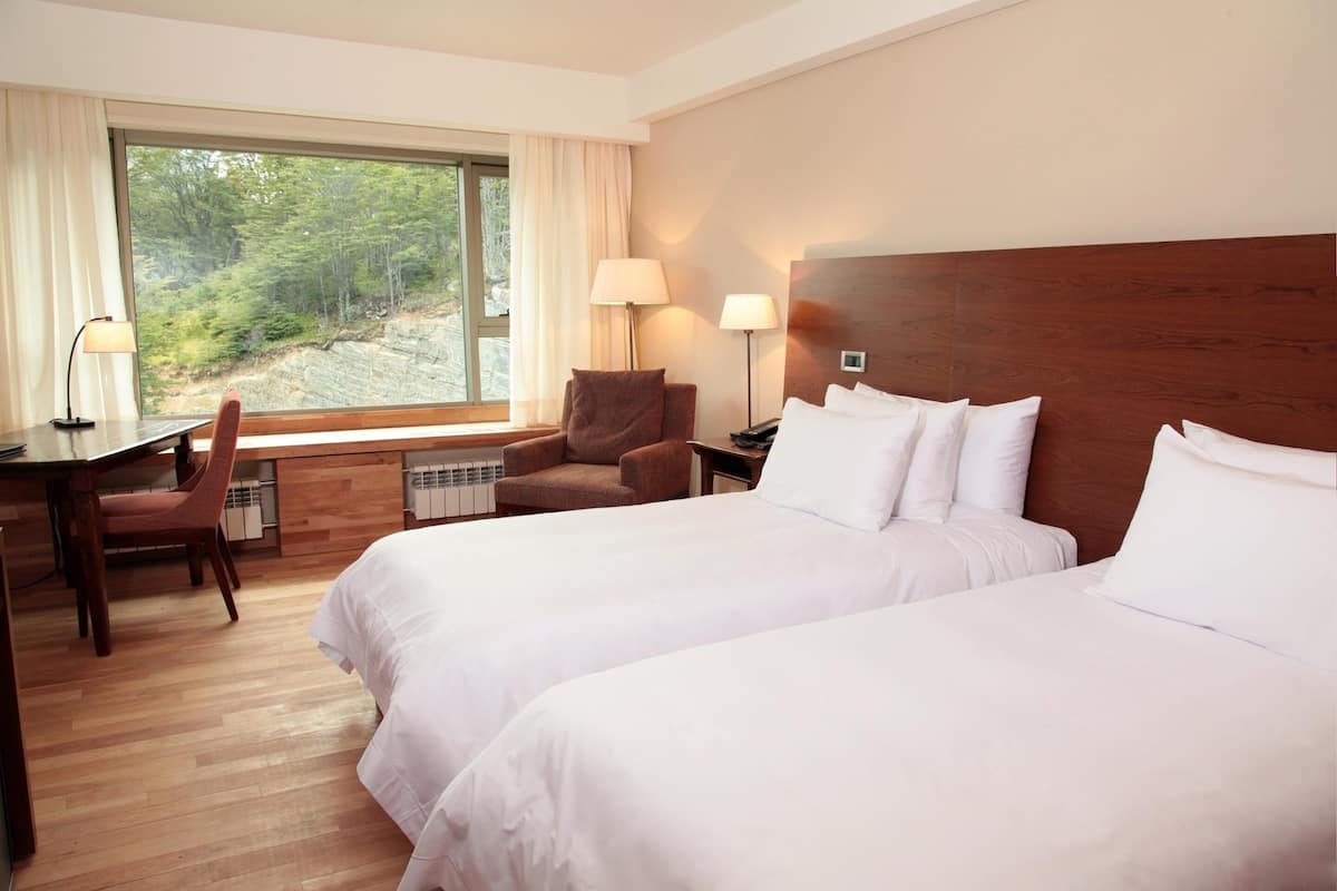 Habitación de hotel con dos camas, un escritorio, una silla y un ventanal con vistas a árboles.