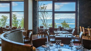 Restaurante con amplios ventanales que ofrecen vistas a montañas y una masa de agua. Las mesas están preparadas con copas de vino y platos.
