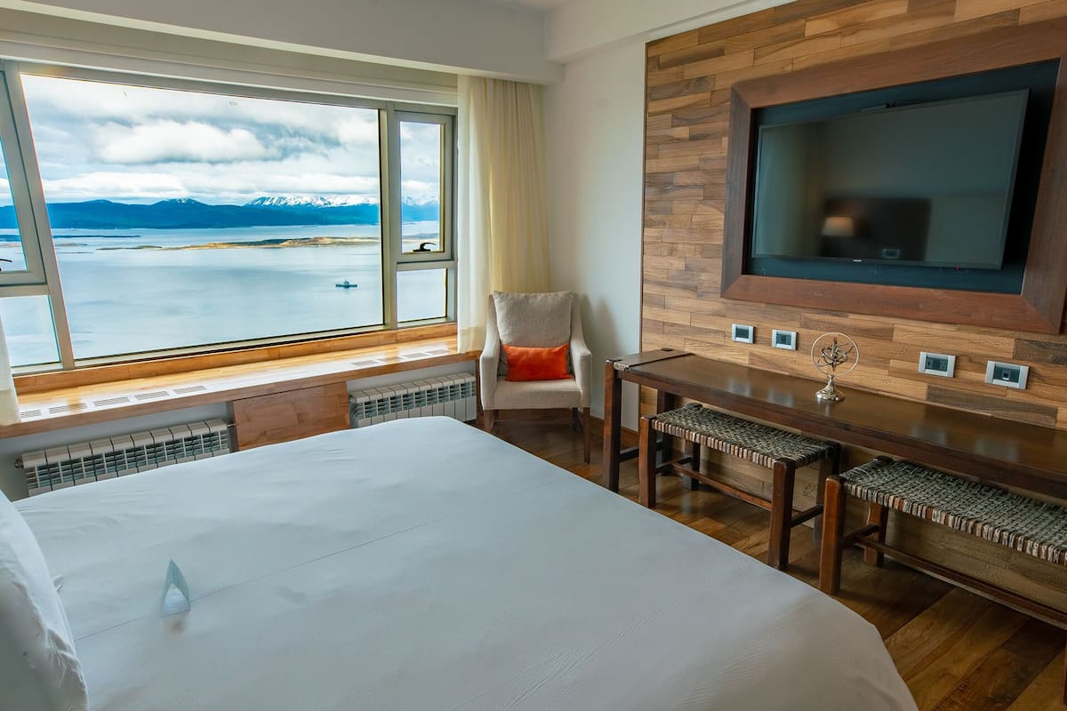 Habitación de hotel con una cama grande, una mesa de comedor, una silla, un televisor montado en una pared de madera y una ventana con vistas a montañas y un lago.