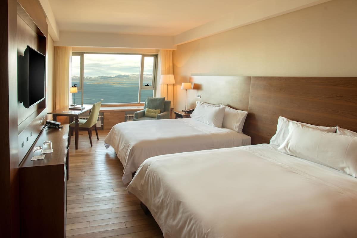 Habitación de hotel con dos camas, un escritorio, una silla, un televisor y una ventana con vista al mar.