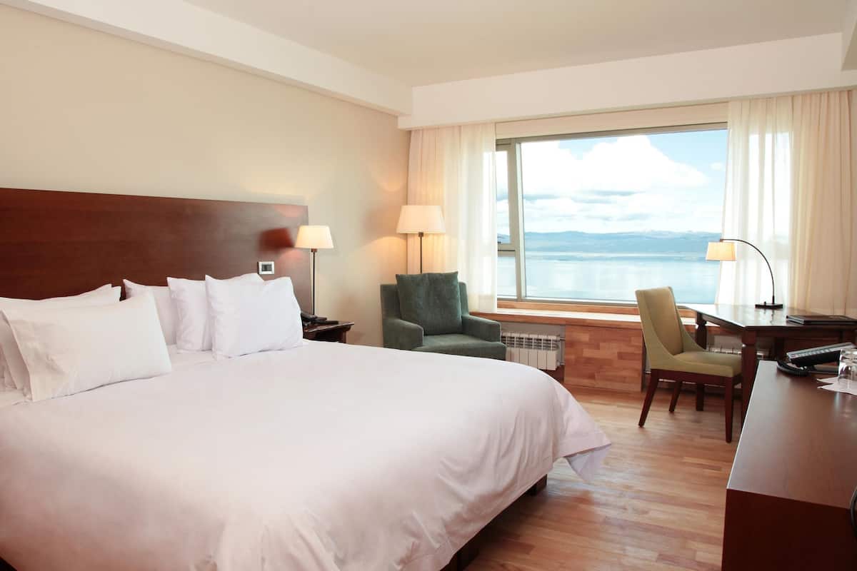 Habitación de hotel con una cama grande, un escritorio con silla, vista al agua y una ventana con cortinas.