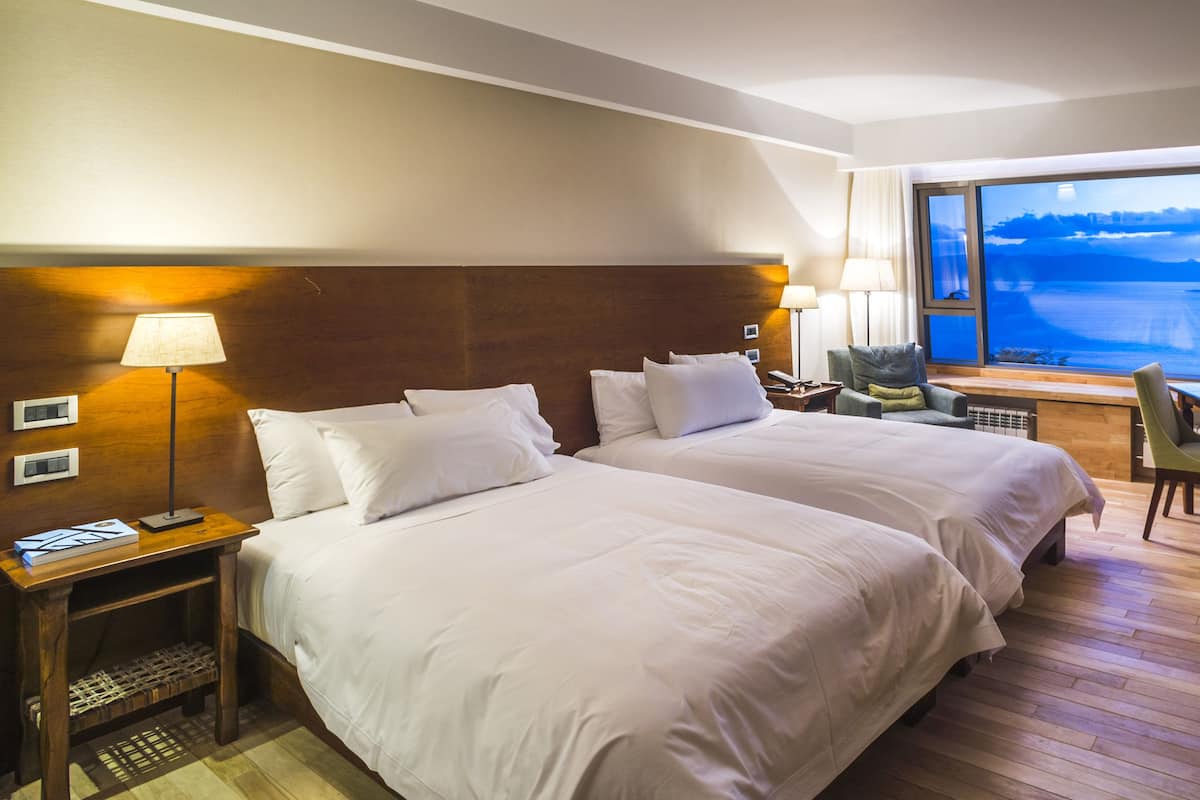 Habitación de hotel con dos camas, un escritorio, una silla y un amplio ventanal con vista a una masa de agua.