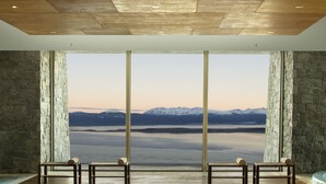 Habitación con un ventanal grande que ofrece vistas a montañas y una masa de agua. Hay dos bancos de madera con cojines.