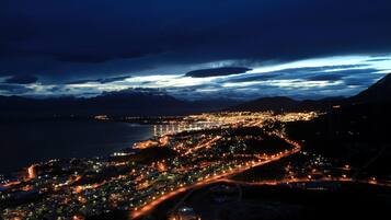 A nighttime aerial view of a coastal city with a prominent lighthouse, winding roads, and mountainous backdrop.