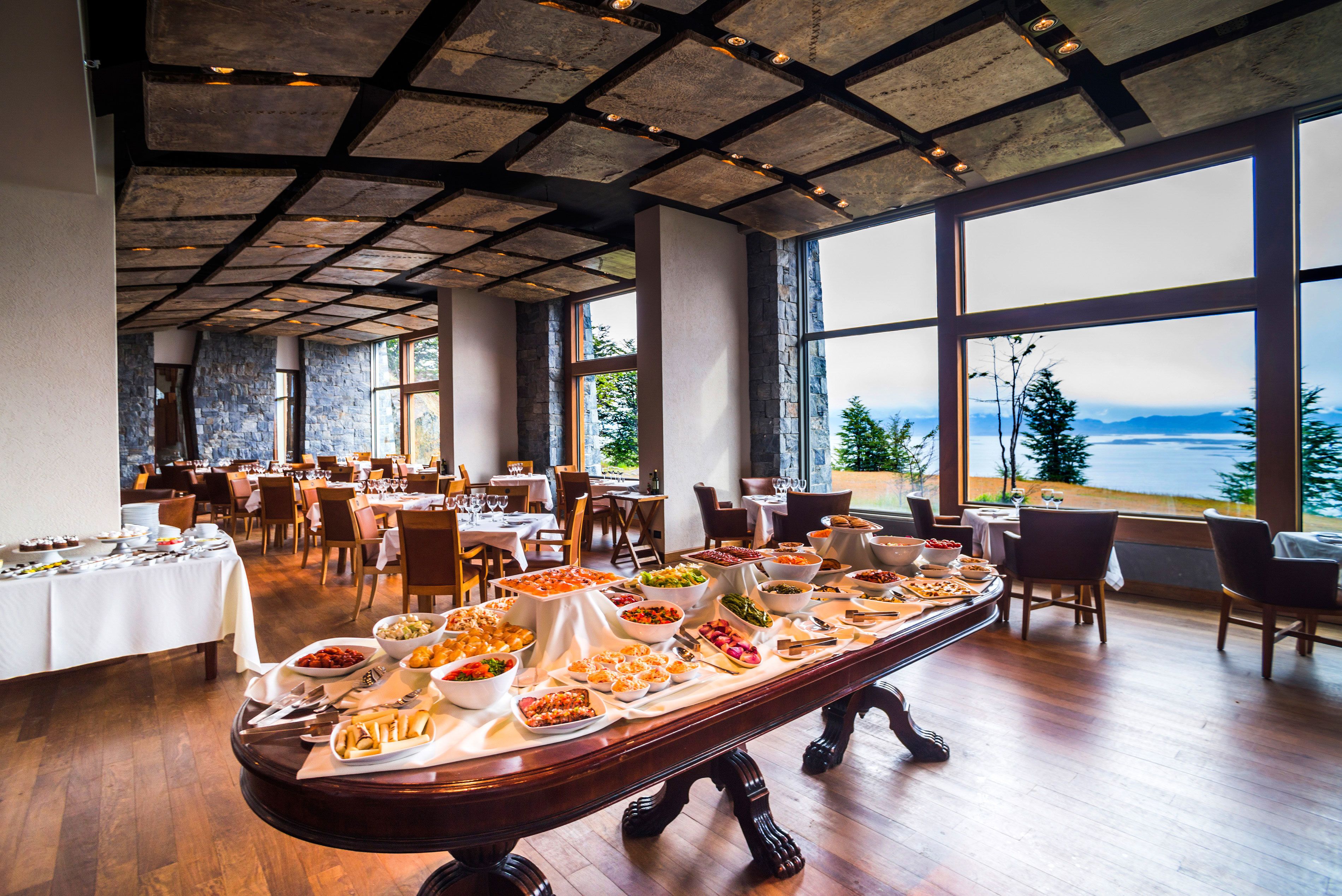 A buffet setup with a variety of dishes, large windows with a view of the outdoors, and a high ceiling with exposed beams.