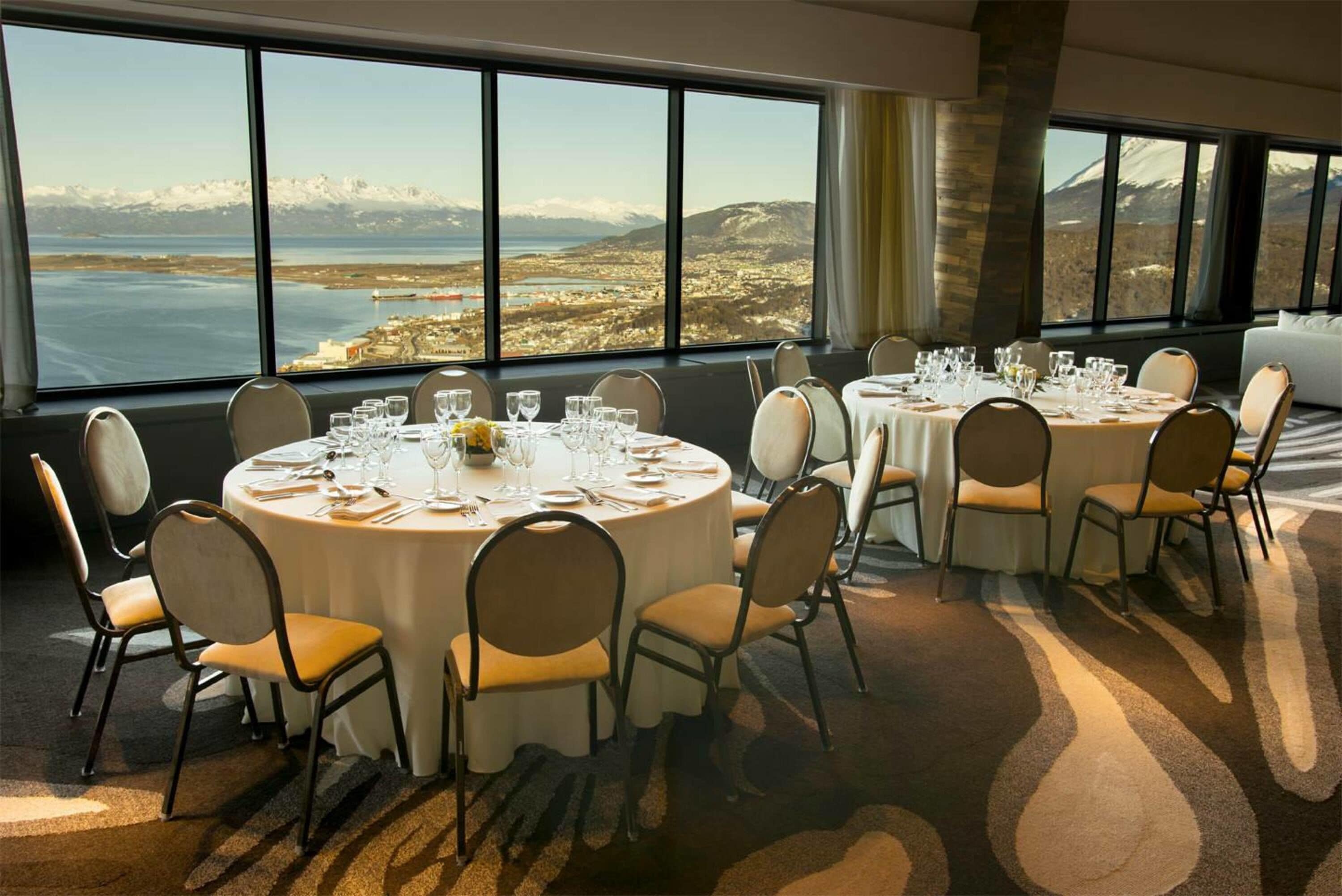 A dining area with round tables set for a meal, large windows offering a view of a mountainous landscape and a body of water.